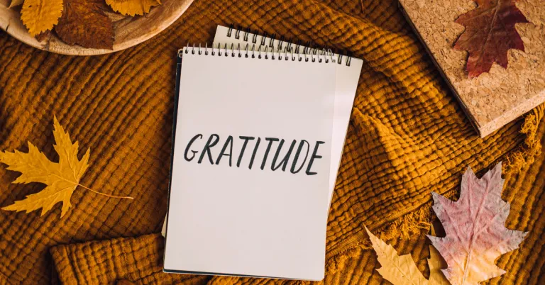 Notebook with the word gratitude written on it, surrounded by fall leaves and a warm autumn blanket.