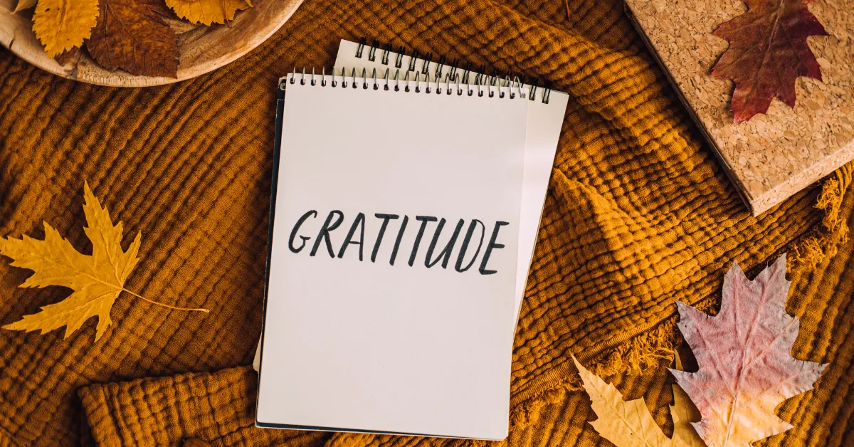 Notebook with the word gratitude written on it, surrounded by fall leaves and a warm autumn blanket.